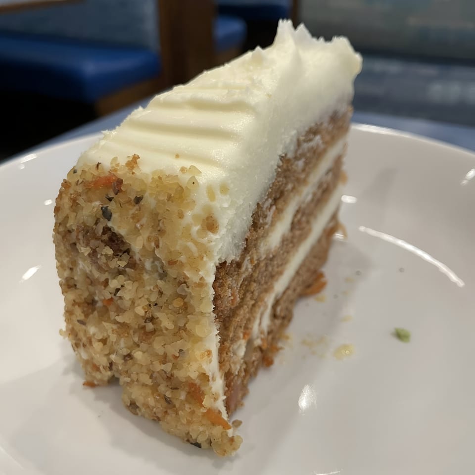 A slice of carrot cake with white frosting and nut topping on a white plate at Sky Diner.