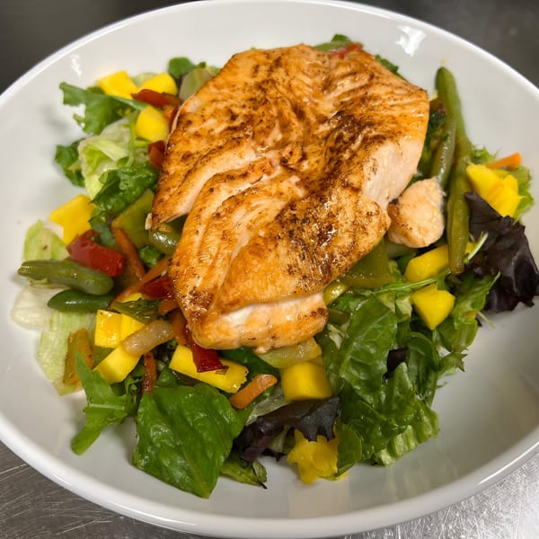 Grilled salmon fillet atop a fresh salad with mango, green beans, and bell peppers in a white bowl.