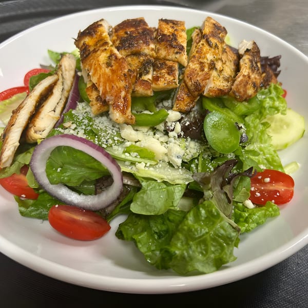 A grilled chicken salad with mixed greens, tomatoes, red onion, and crumbled cheese in a white bowl.