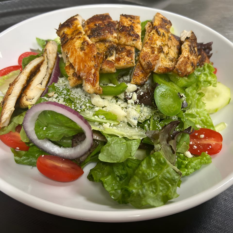 A grilled chicken salad with mixed greens, tomatoes, red onion, and crumbled cheese in a white bowl.