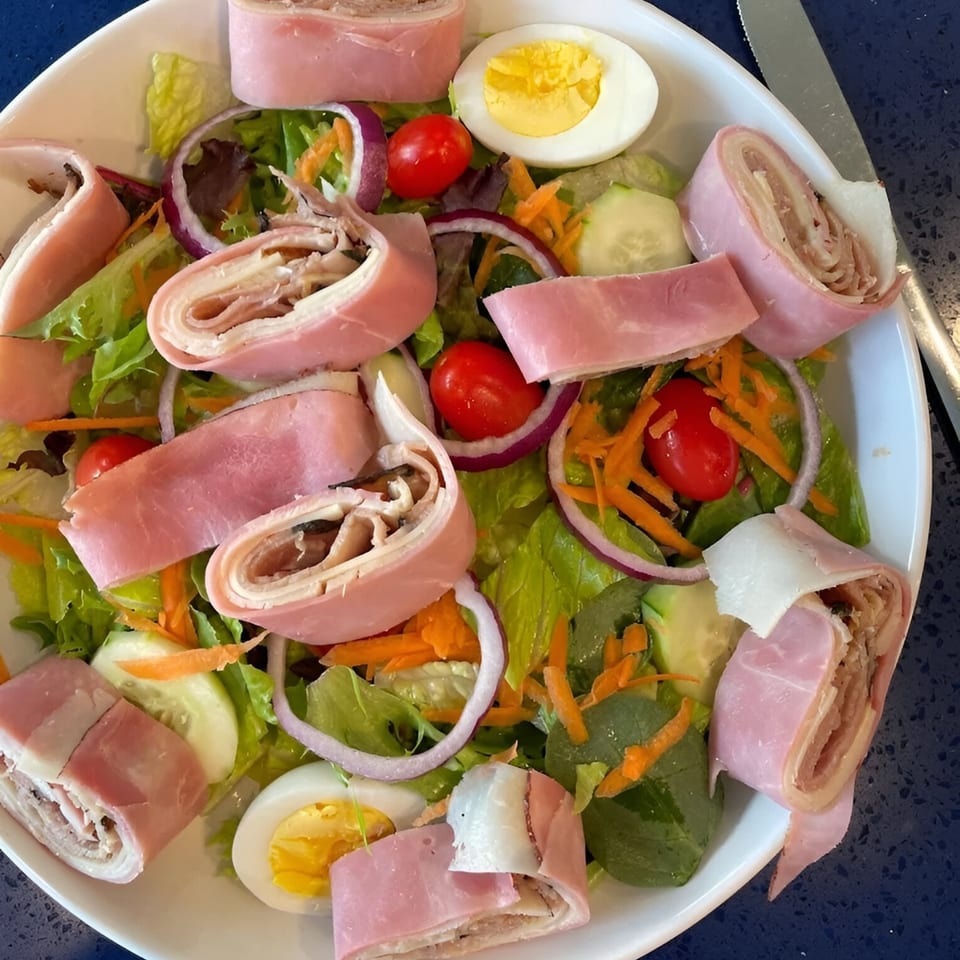 A chef salad with ham and cheese roll-ups, mixed greens, tomatoes, red onion, and hard-boiled egg.