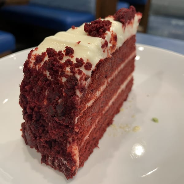 A slice of red velvet cake with white cream cheese frosting and red crumbs on a white plate.